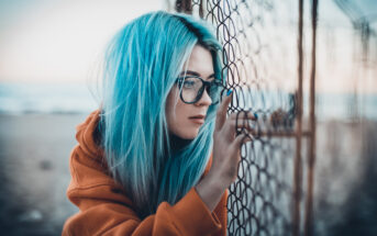 A person with long, vibrant blue hair and glasses is standing beside a wire fence, gazing thoughtfully through it. They are wearing an orange hoodie and appear to be near a beach,with the sea and sky visible in the background.