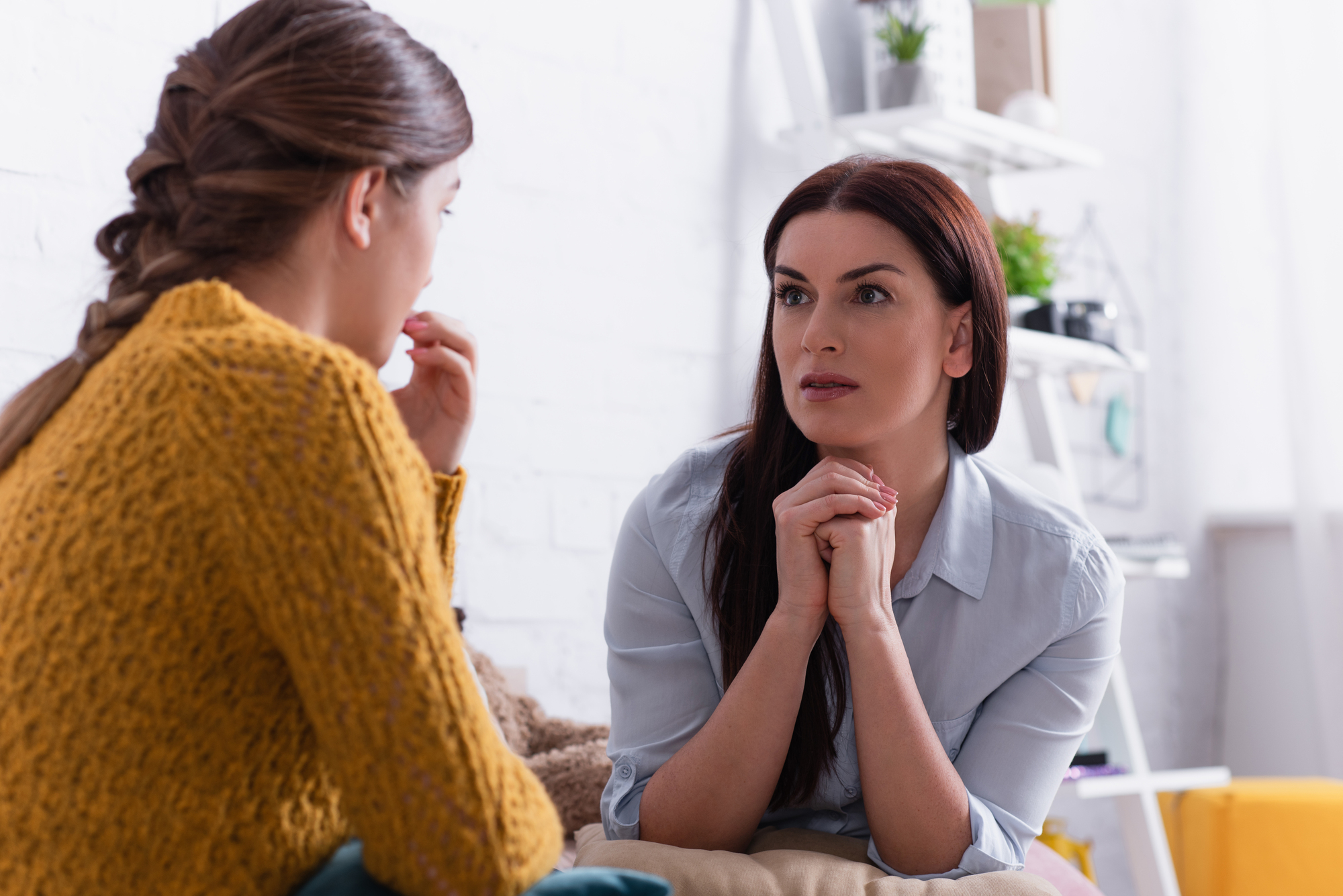 Two people are sitting indoors, having a serious conversation. One has a worried expression and is wearing a yellow sweater, while the other listens intently with hands clasped, dressed in a light blue shirt. The setting appears cozy with some plants and decor in the background.