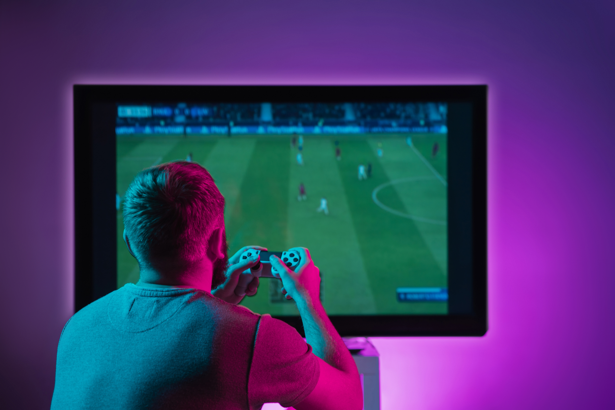 A person with short hair, seen from behind, is holding a video game controller and playing a soccer video game on a large screen. The room is dimly lit with a pink and blue glow, illuminating the player and the screen.