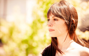 Profile view of a young woman with long, straight brown hair and freckles, standing outdoors on a sunny day. The background is blurred with bright green foliage and sunlight, highlighting her contemplative expression.