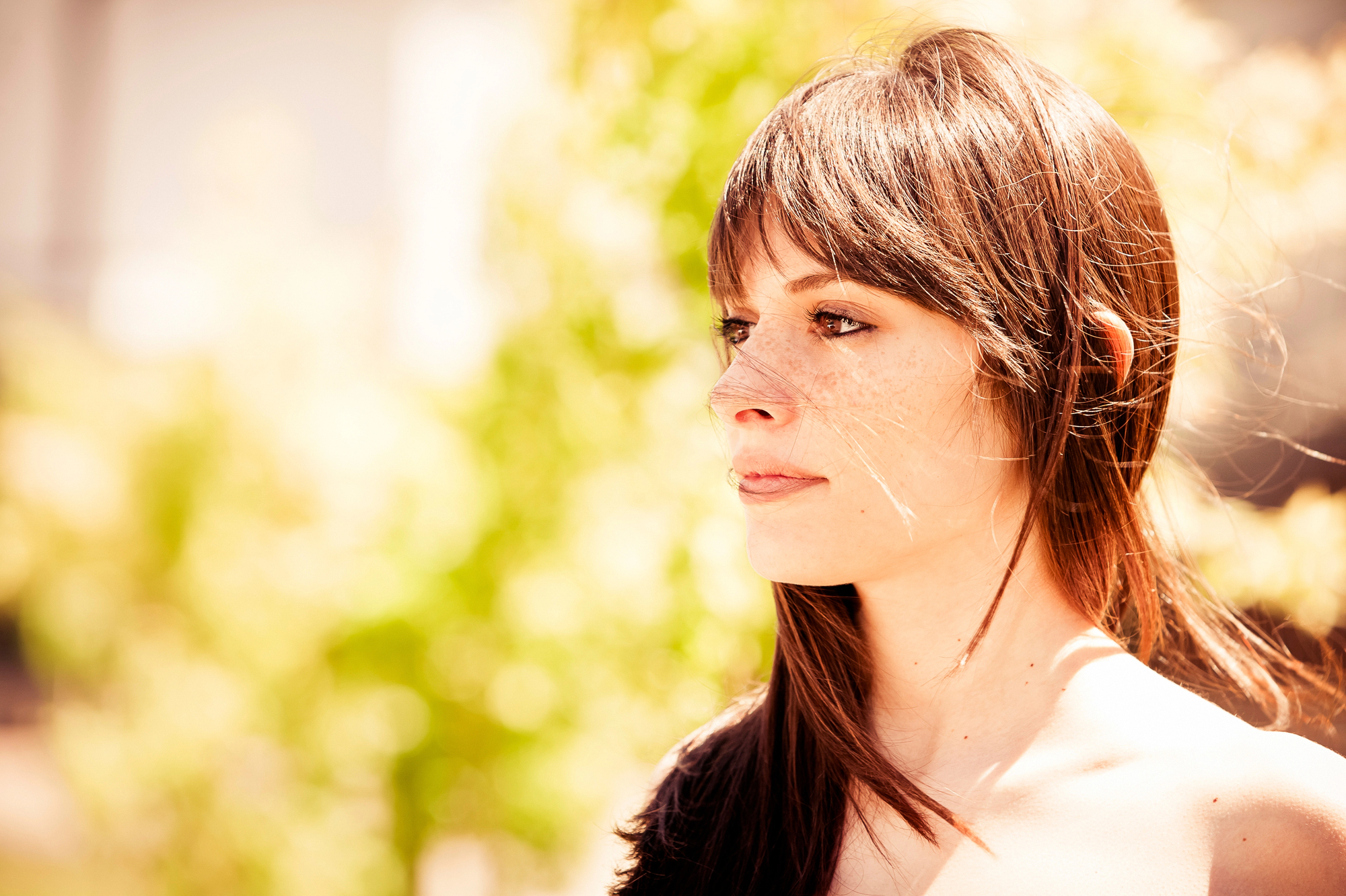 Profile view of a young woman with long, straight brown hair and freckles, standing outdoors on a sunny day. The background is blurred with bright green foliage and sunlight, highlighting her contemplative expression.