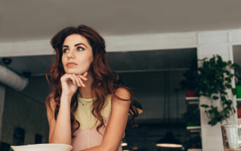 A woman with long, wavy brown hair wearing a beige and pink sleeveless top is sitting at a table. She is resting her chin on her hand and gazing thoughtfully to the side. The background features indoor plants and a softly lit, modern interior.