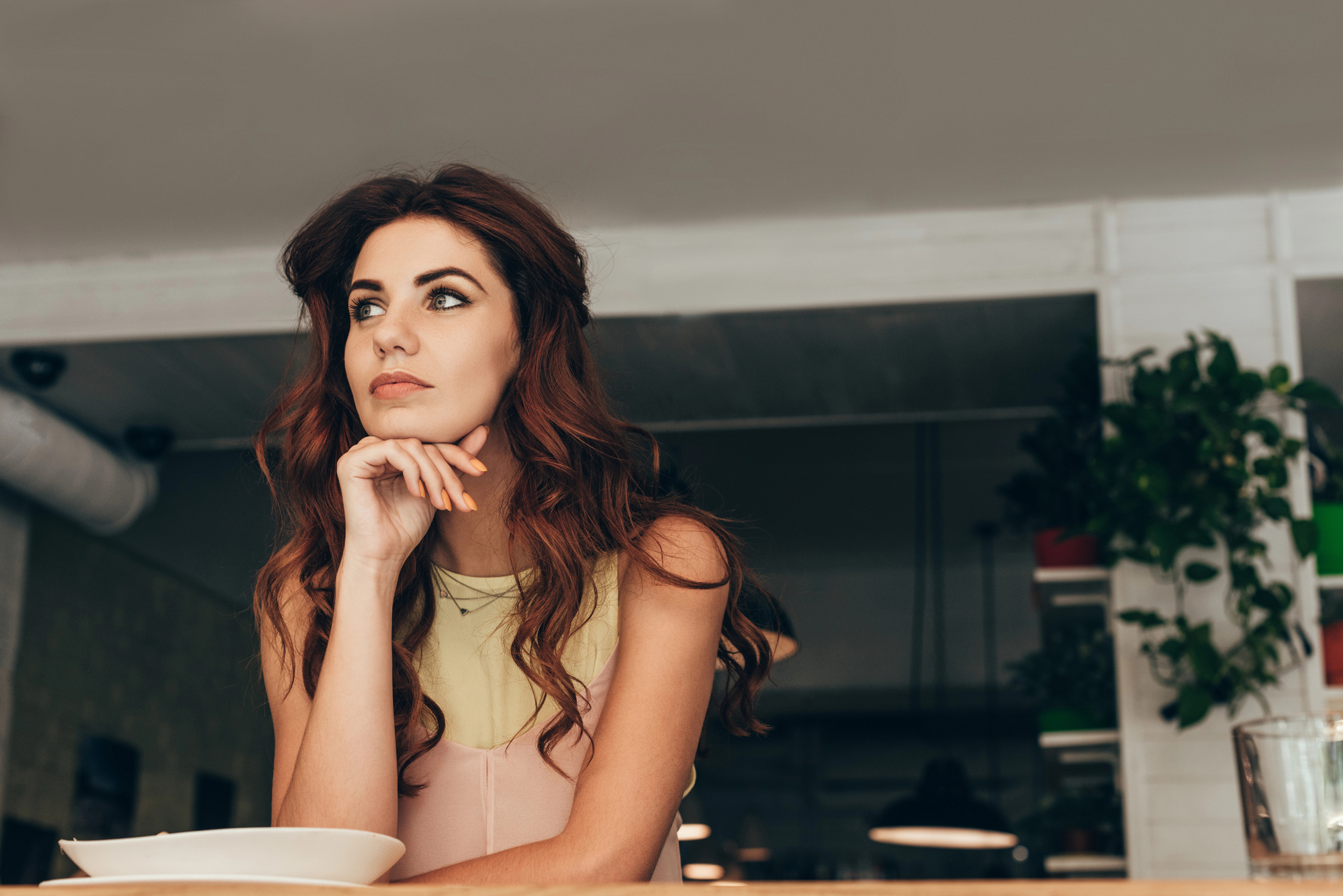 A woman with long, wavy brown hair wearing a beige and pink sleeveless top is sitting at a table. She is resting her chin on her hand and gazing thoughtfully to the side. The background features indoor plants and a softly lit, modern interior.