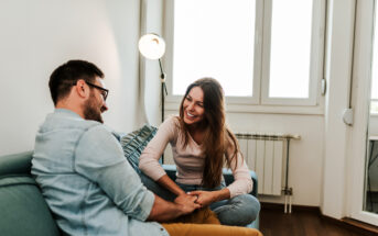 A man and a woman sit on a couch in a bright living room, smiling and holding hands. They appear to be engaged in a cheerful conversation. The room has white walls, a floor lamp, and large windows allowing natural light to fill the space.