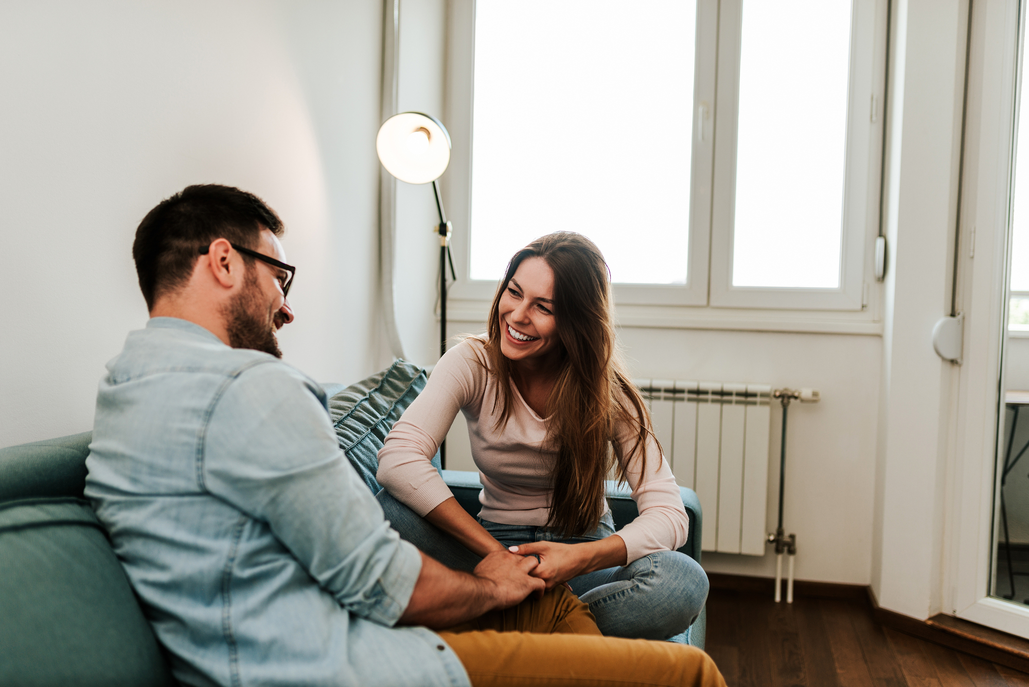 A man and a woman sit on a couch in a bright living room, smiling and holding hands. They appear to be engaged in a cheerful conversation. The room has white walls, a floor lamp, and large windows allowing natural light to fill the space.