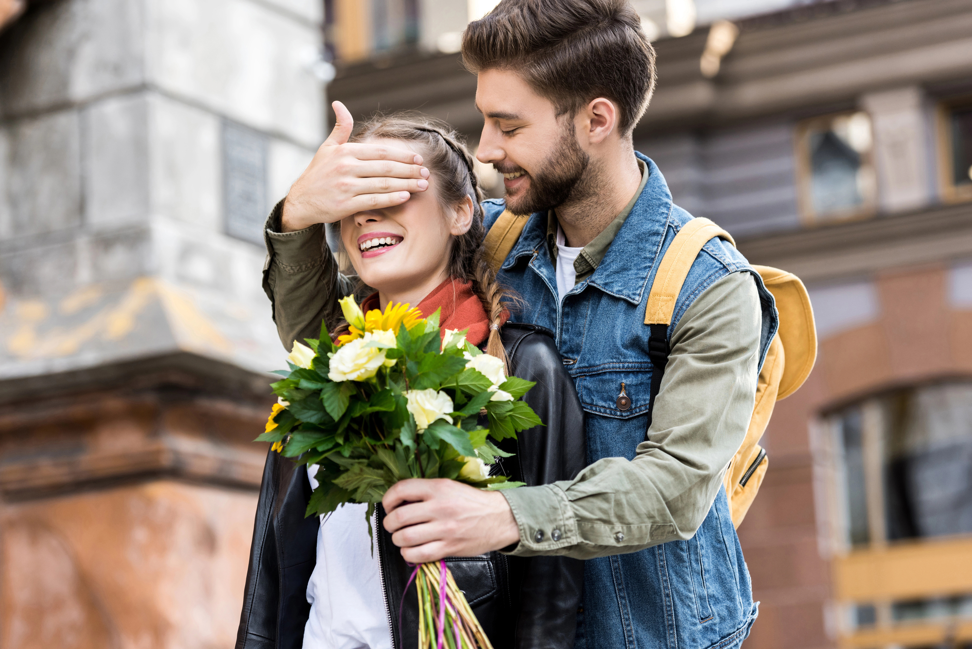 A smiling man with a beard in a denim jacket playfully covers the eyes of a woman holding a bouquet of flowers. They are both standing outdoors in an urban setting, with buildings in the background. The woman is laughing and wearing a leather jacket.