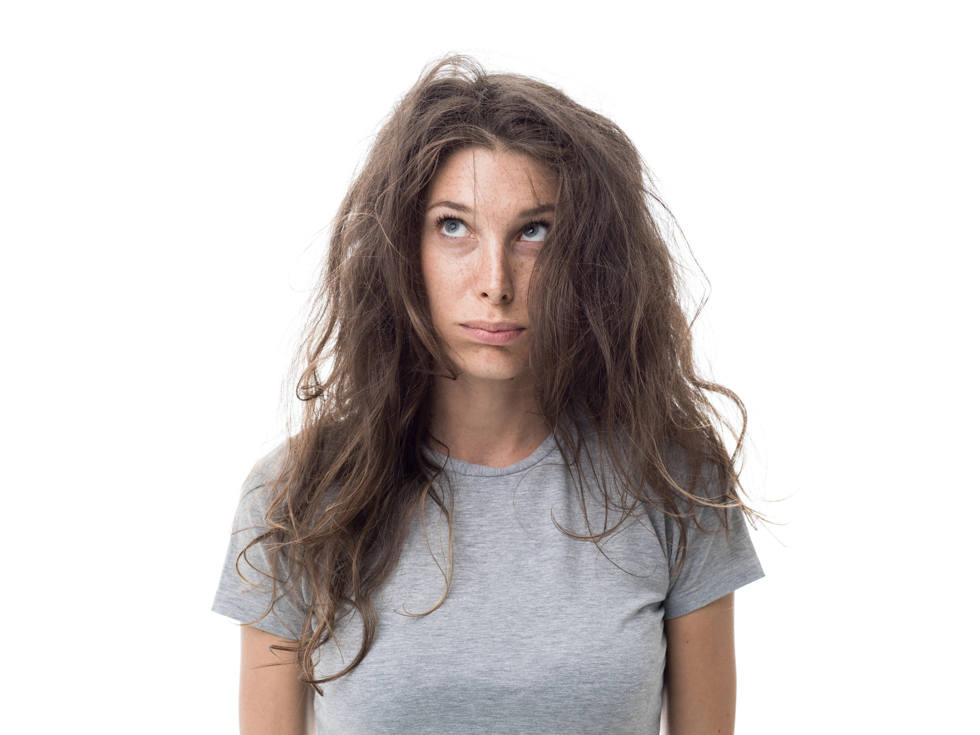 A woman with long, messy hair wearing a light gray T-shirt looks upwards with a slightly puzzled or frustrated expression against a plain white background.