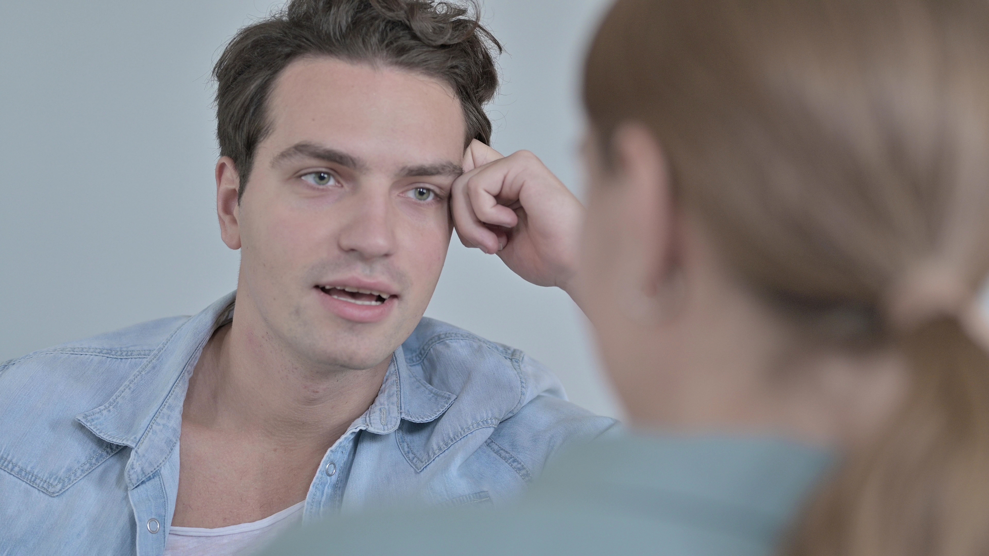 A man with dark hair, wearing a light blue denim shirt, is engaged in a conversation with a woman whose back is to the camera. The man rests his head on his hand and looks attentively at the woman in a casual indoor setting.