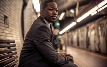 A man in a dark suit sits on a bench at a subway station. He looks to his right, seemingly deep in thought. The subway platform features white tiles, and there is a train on the opposite track. The station is dimly lit, with lights creating a warm ambiance.