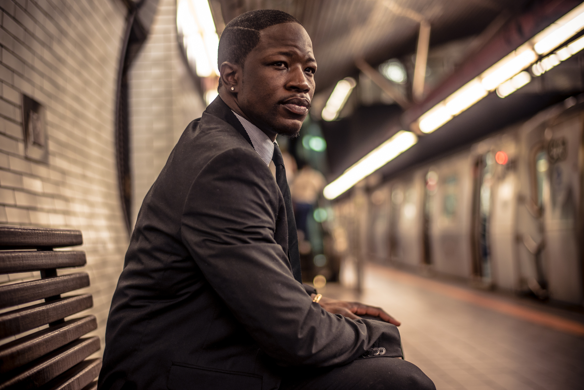 A man in a dark suit sits on a bench at a subway station. He looks to his right, seemingly deep in thought. The subway platform features white tiles, and there is a train on the opposite track. The station is dimly lit, with lights creating a warm ambiance.