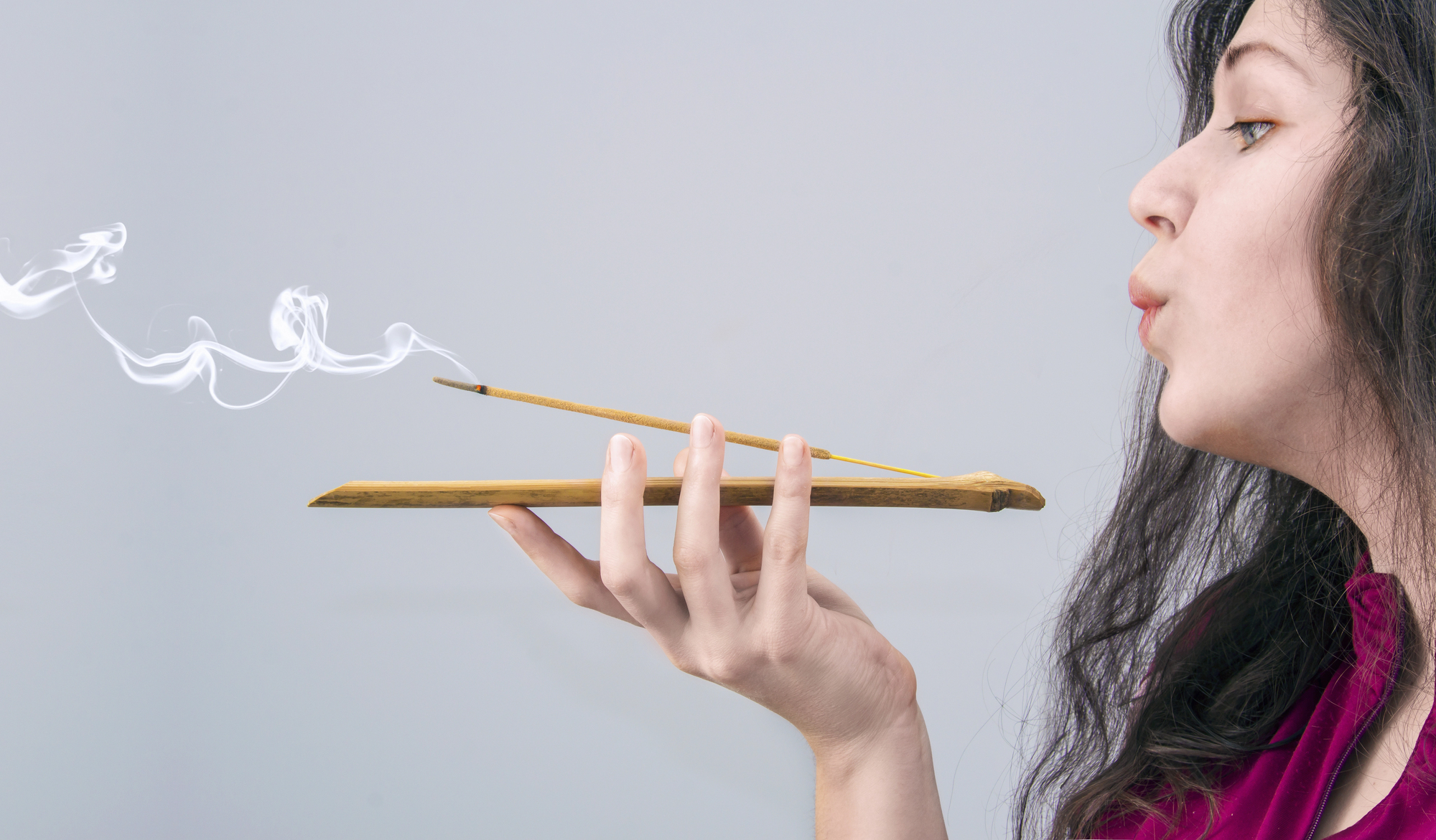 A woman blows on two smoking incense sticks placed on a wooden holder that she is holding with one hand. The background is plain and gray. The woman has long dark hair and is wearing a pink top.