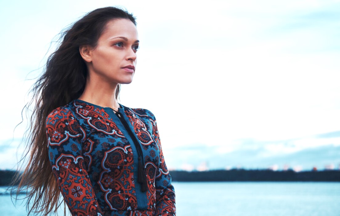 A woman stands in front of a large body of water, gazing into the distance. She has long, flowing hair and is wearing a patterned, long-sleeve dress with intricate designs in various colors. The background shows a serene, slightly cloudy sky.