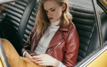 A woman with blonde hair sits in the back seat of a car, wearing a red leather jacket, white turtleneck, and yellow pants. She is looking down and using a smartphone. The interior of the car is black with textured upholstery.