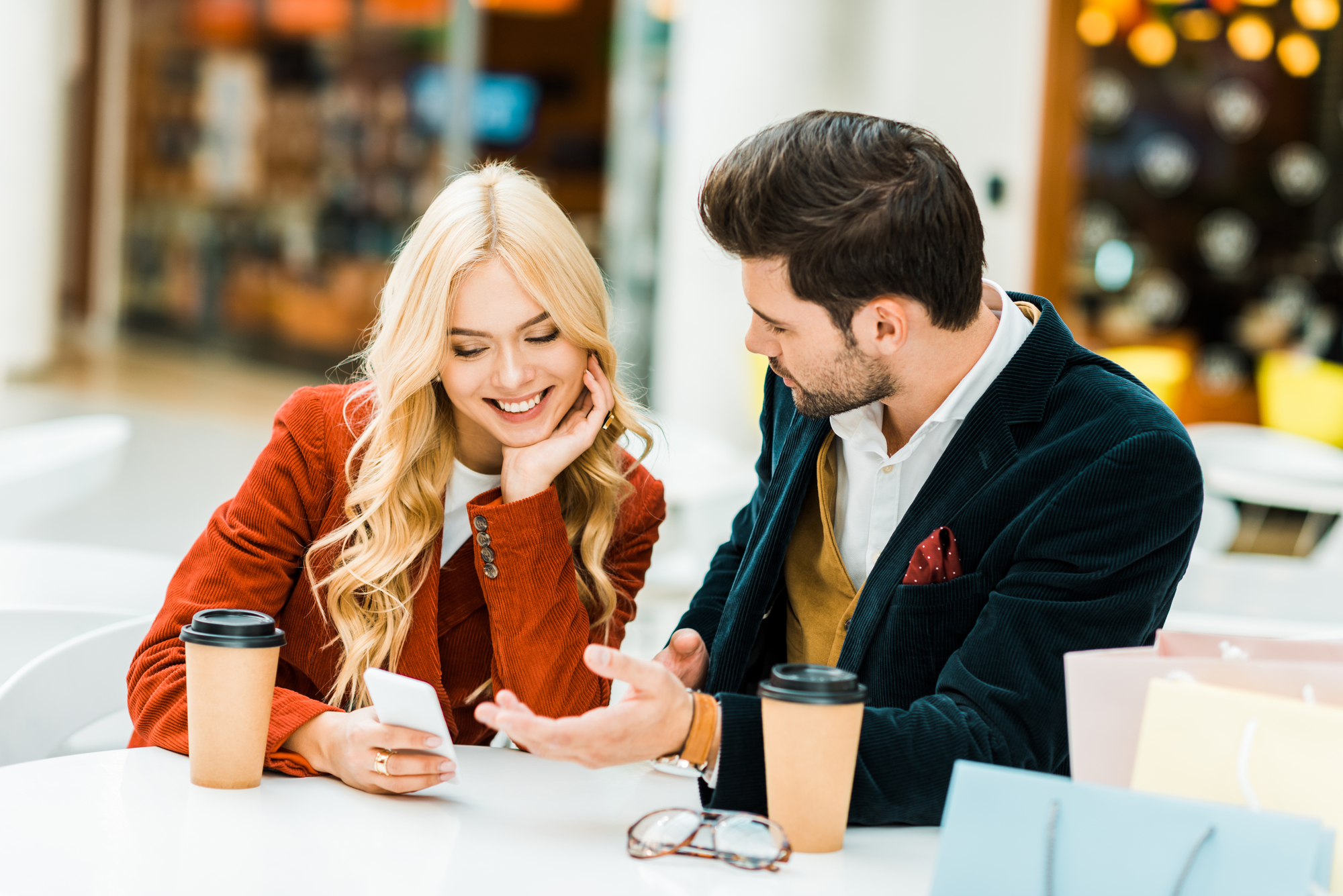 A woman and a man sit at a table in a shopping mall, smiling while looking at a smartphone. Both have takeaway coffee cups on the table. The woman has long blonde hair and wears a red jacket, and the man has short dark hair, a dark jacket, and a yellow sweater.