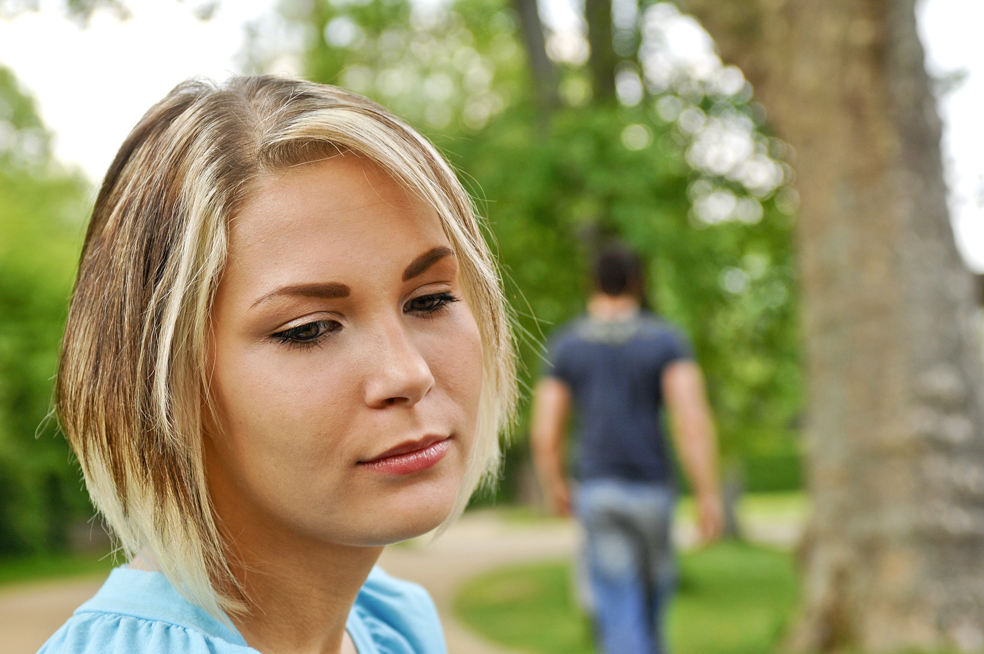 A woman with short blonde hair looks pensive as she gazes downward in a park. In the blurred background, a man walks away on a path, surrounded by trees.