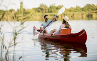 A man and a woman paddle a red canoe on a calm lake surrounded by greenery. The man, in a striped shirt and sunglasses, smiles at the woman, who is wearing a white hat and a pink shirt. Both appear to be enjoying a sunny, peaceful day on the water.