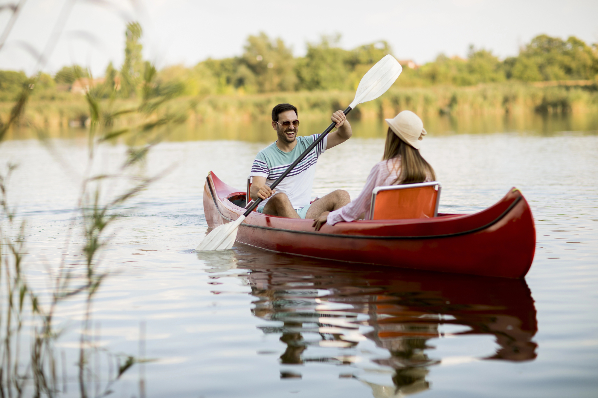 A man and a woman paddle a red canoe on a calm lake surrounded by greenery. The man, in a striped shirt and sunglasses, smiles at the woman, who is wearing a white hat and a pink shirt. Both appear to be enjoying a sunny, peaceful day on the water.