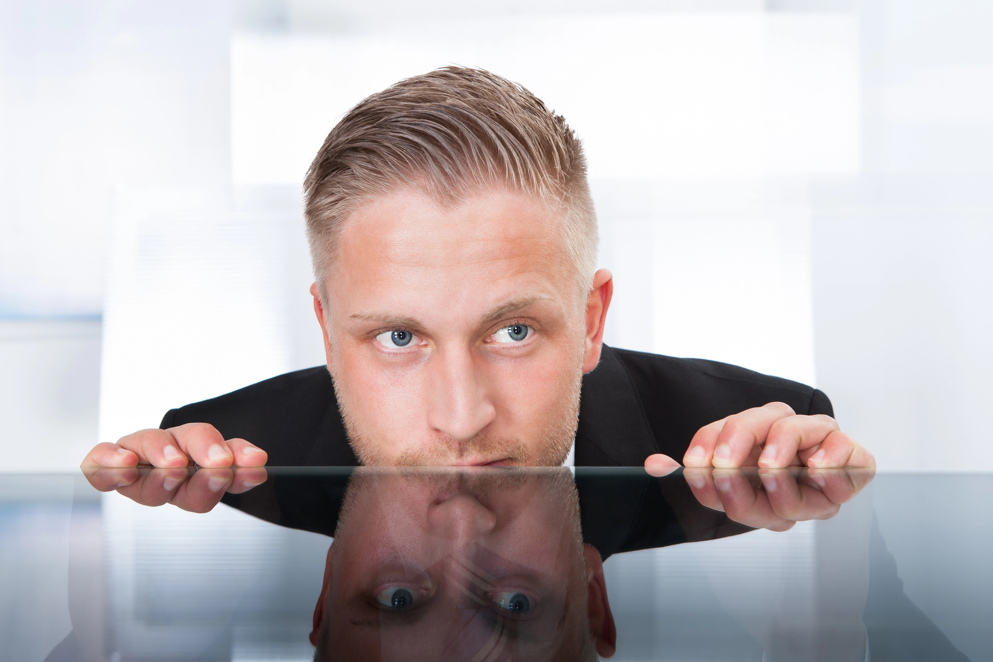 A man with short, light brown hair wearing a black suit peers over a glass table, his reflection visible on the surface. He has an inquisitive expression, and the background is a softly focused, bright indoor setting.