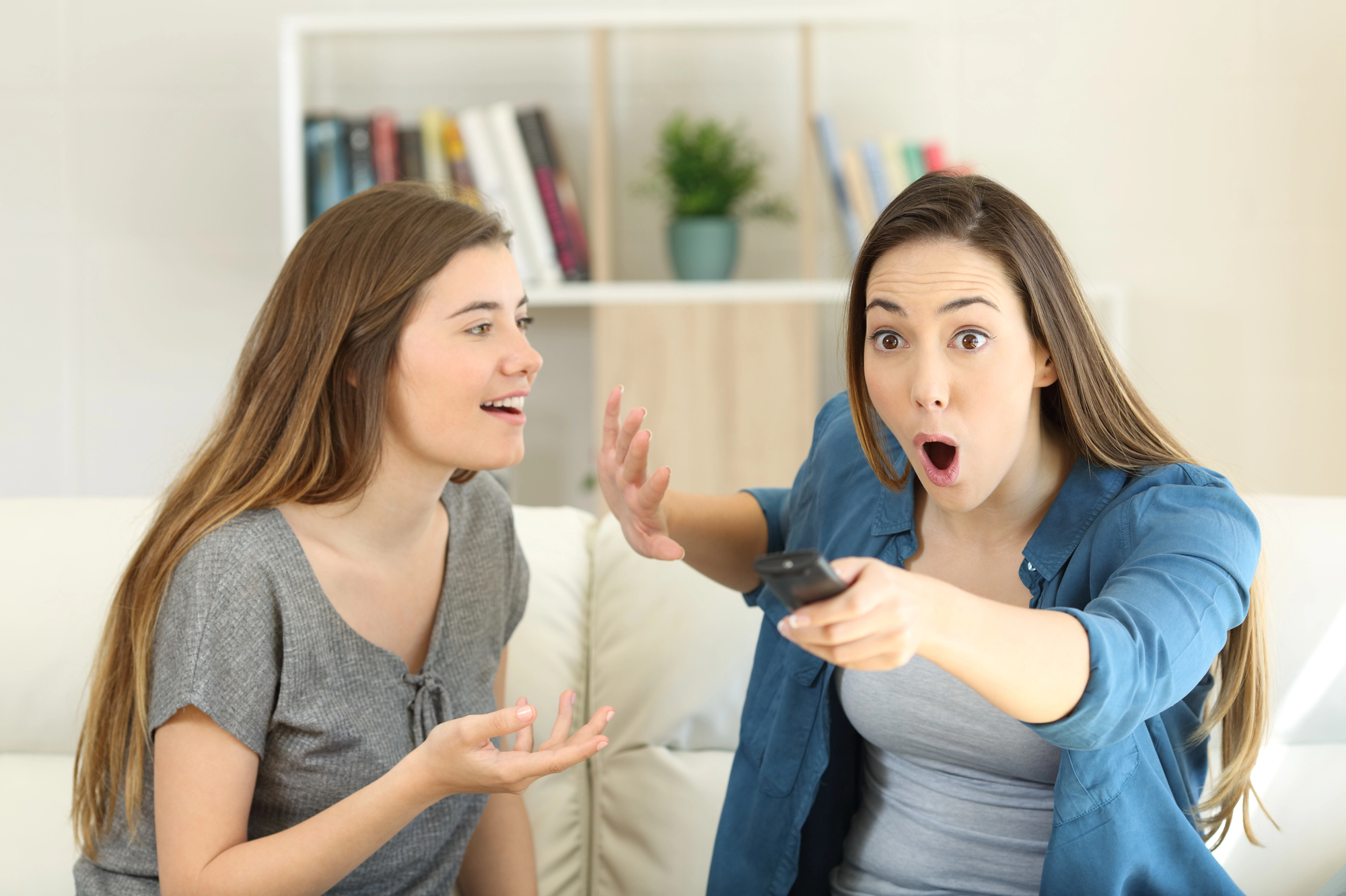 Two young women are sitting on a couch in a living room. One woman with long brown hair, wearing a grey T-shirt, is talking animatedly. The other woman, also with long brown hair and wearing a blue shirt, looks surprised while holding a TV remote. A bookshelf is in the background.