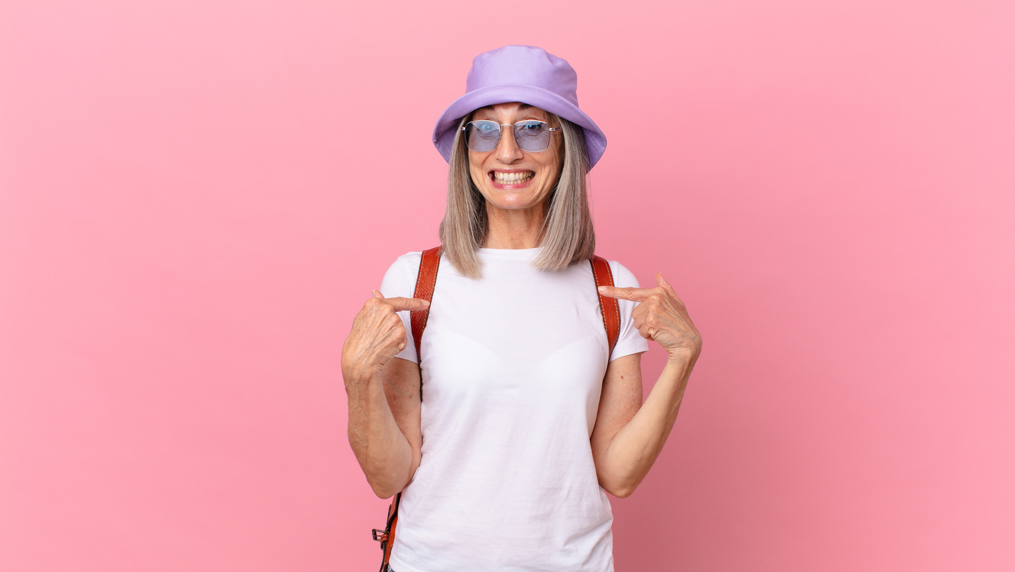 A smiling person with gray hair wearing a light purple bucket hat, eyeglasses, a white t-shirt, and a red backpack stands against a pink background, pointing to their t-shirt with both hands.