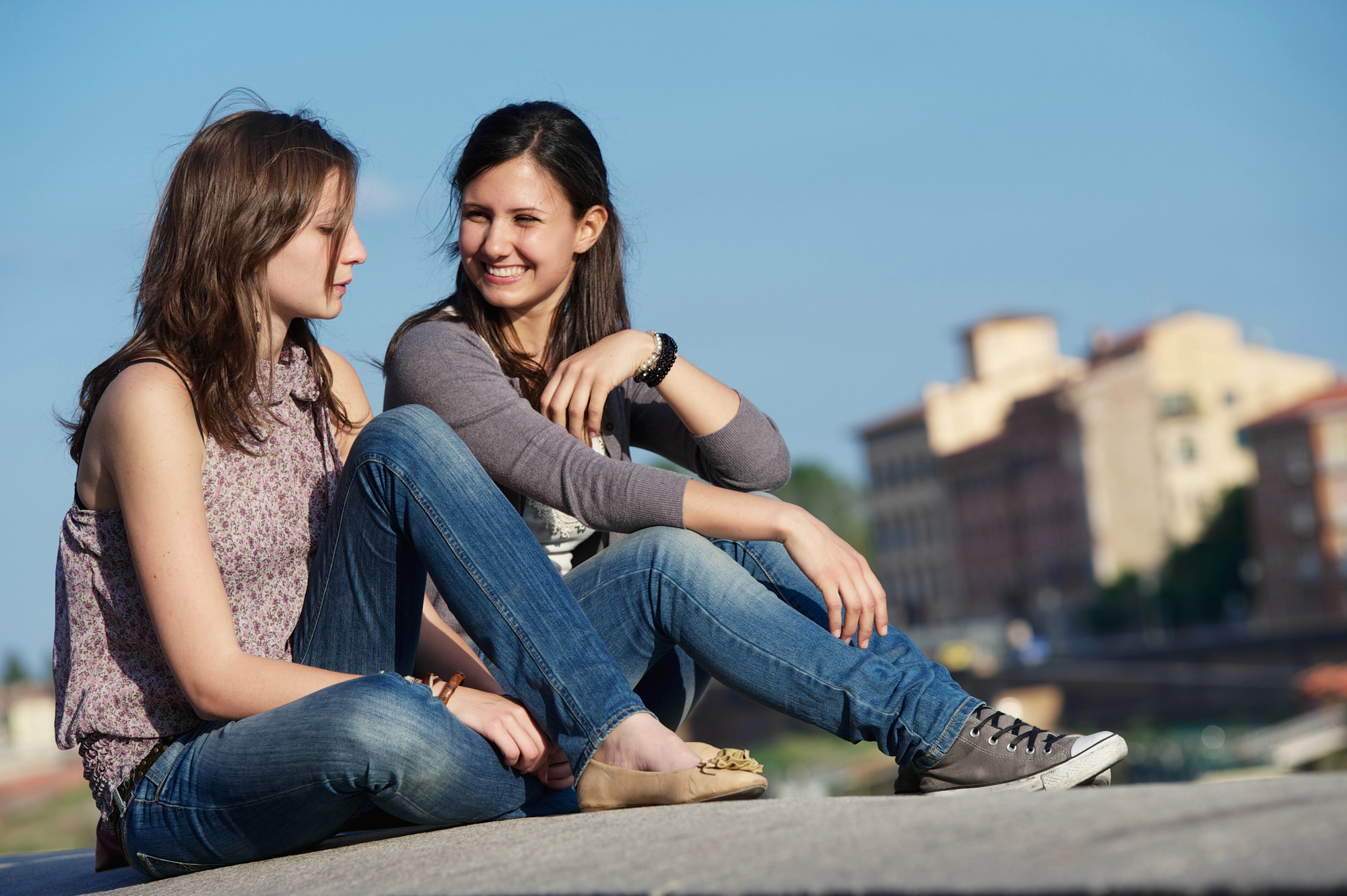 Two young women sit casually on a concrete ledge outdoors. Both are dressed in jeans and casual tops, engaged in a relaxed conversation. The background features buildings and a blue sky, suggesting an urban setting. One woman is smiling while the other listens attentively.