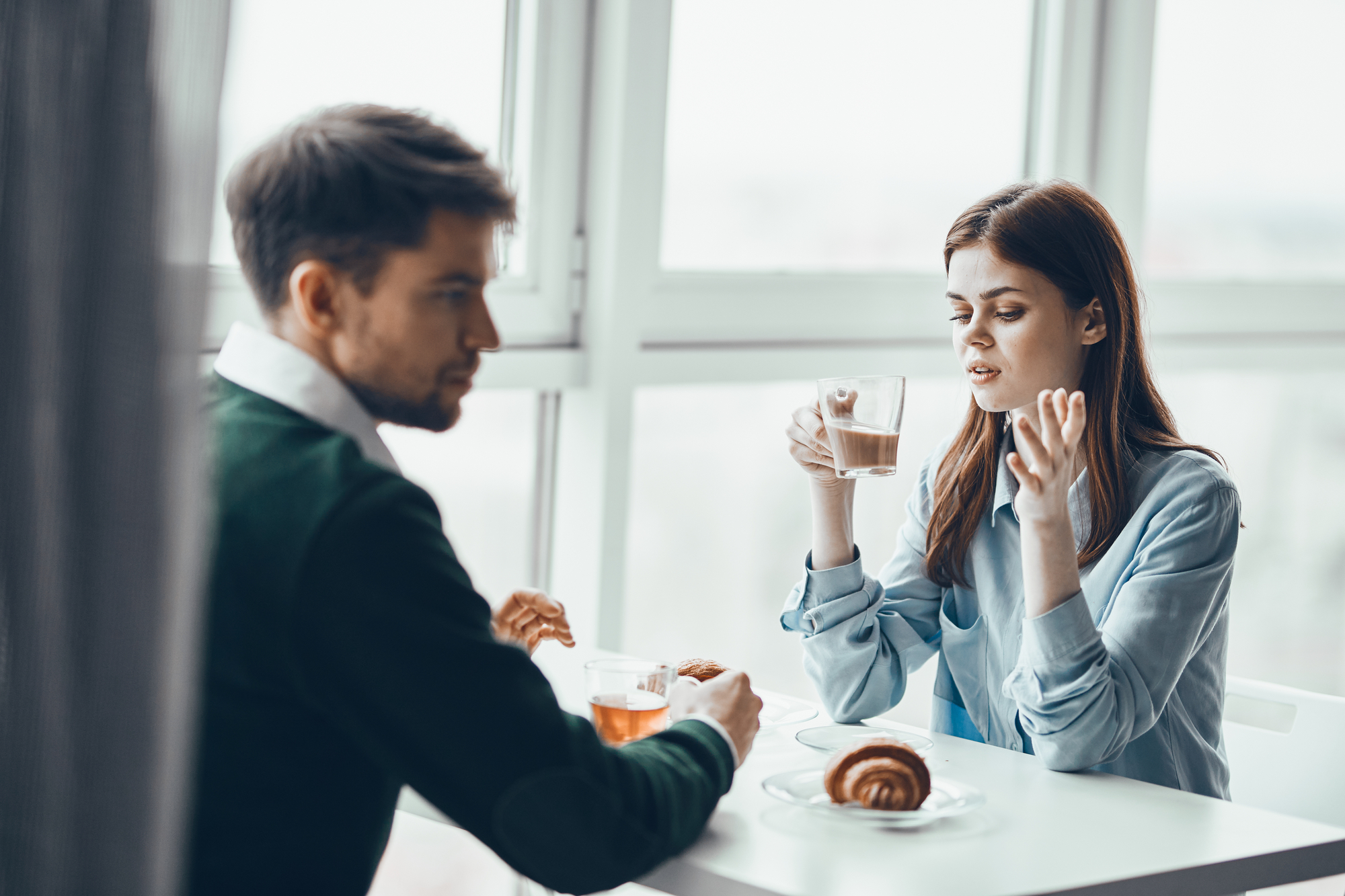 A man and woman are sitting at a table by a window, having a conversation. The woman gestures with one hand while holding a glass with a drink in the other. There are croissants and cups on the table. The setting appears to be a bright, sunny room.