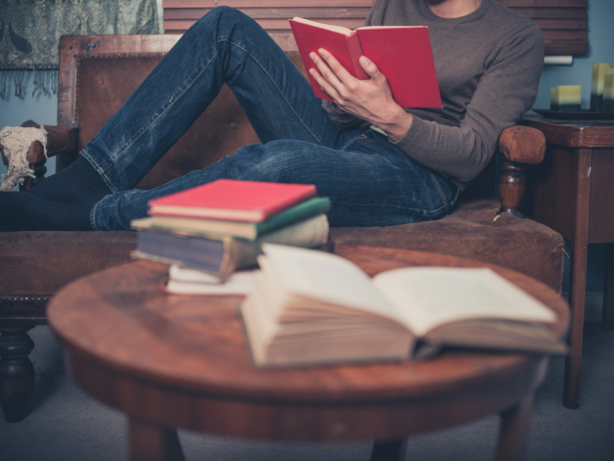 A person is reading a red book while reclining on a brown couch. They are wearing jeans and a brown sweater. In the foreground, there is a wooden round table with several books, one of which is open. The room has a cozy and relaxed atmosphere.