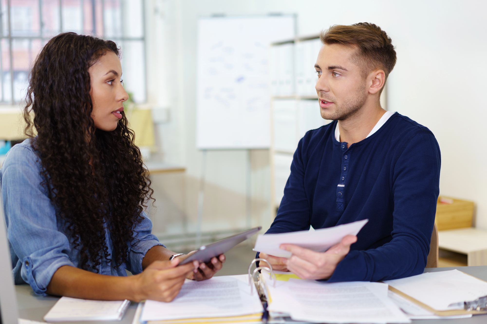 A woman holding a tablet and a man holding documents are engaged in a discussion at a desk filled with papers. A whiteboard with notes stands in the background in a well-lit office environment.