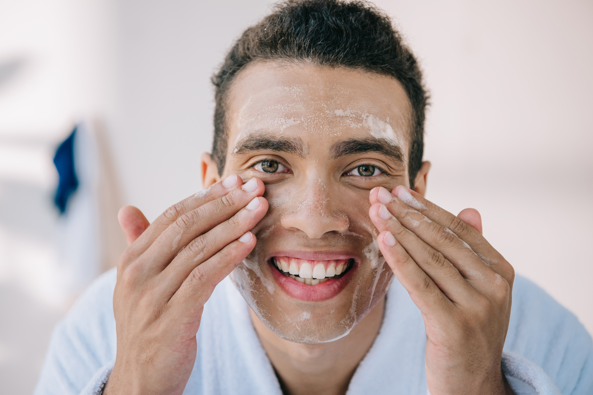 A person with a bright smile applies a skincare product to their face using both hands. They are wearing a light-colored bathrobe and appear to be in a bathroom, engaging in a morning skincare routine. The background is softly blurred.