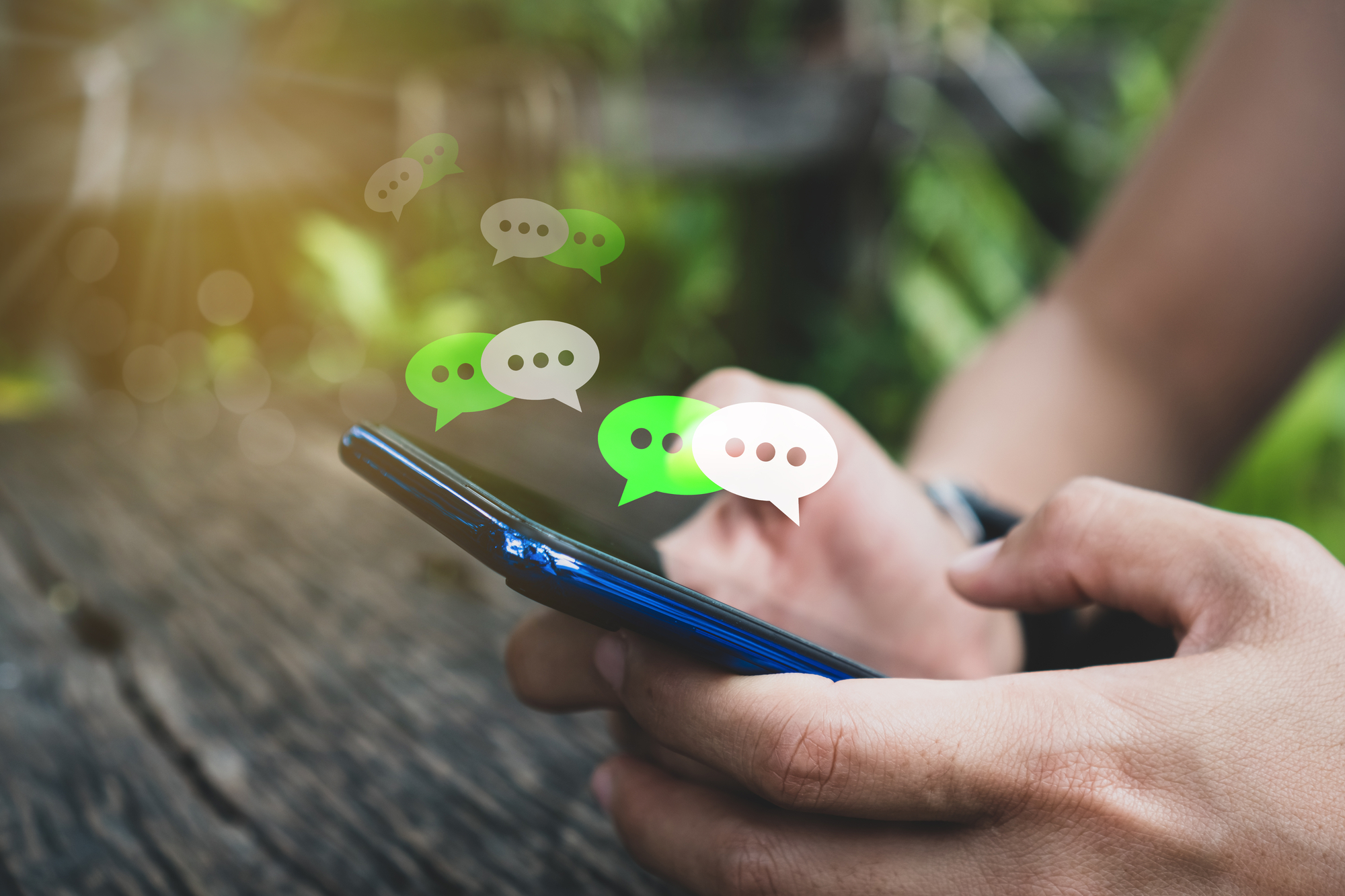 A person holds a smartphone with both hands, appearing engaged in a messaging conversation. Several speech bubbles in green and white are floating above the phone, symbolizing ongoing text messages. The background is blurred with greenery.