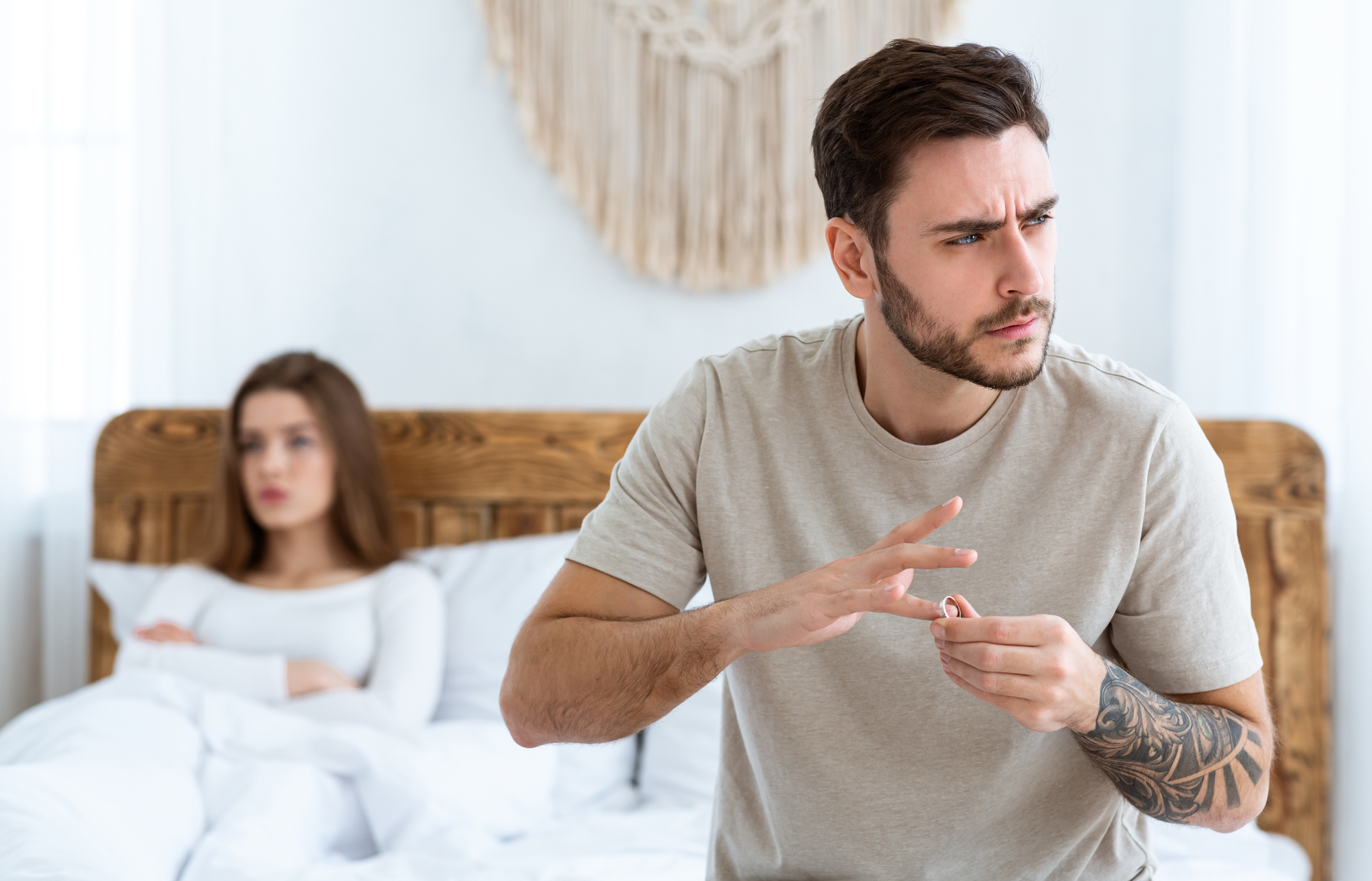 A man with a beard and tattoos, dressed in a beige t-shirt, is taking off his wedding ring while looking away with a concerned expression. He stands in front of a bed where a woman with long brown hair, dressed in white, sits with a sad expression.