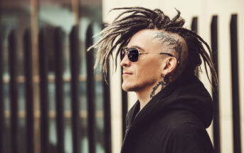 A person with a shaved head and long dreadlocks on top is wearing sunglasses and a black jacket. They have tattoos on their head and face. The background is blurred, featuring vertical architectural elements.