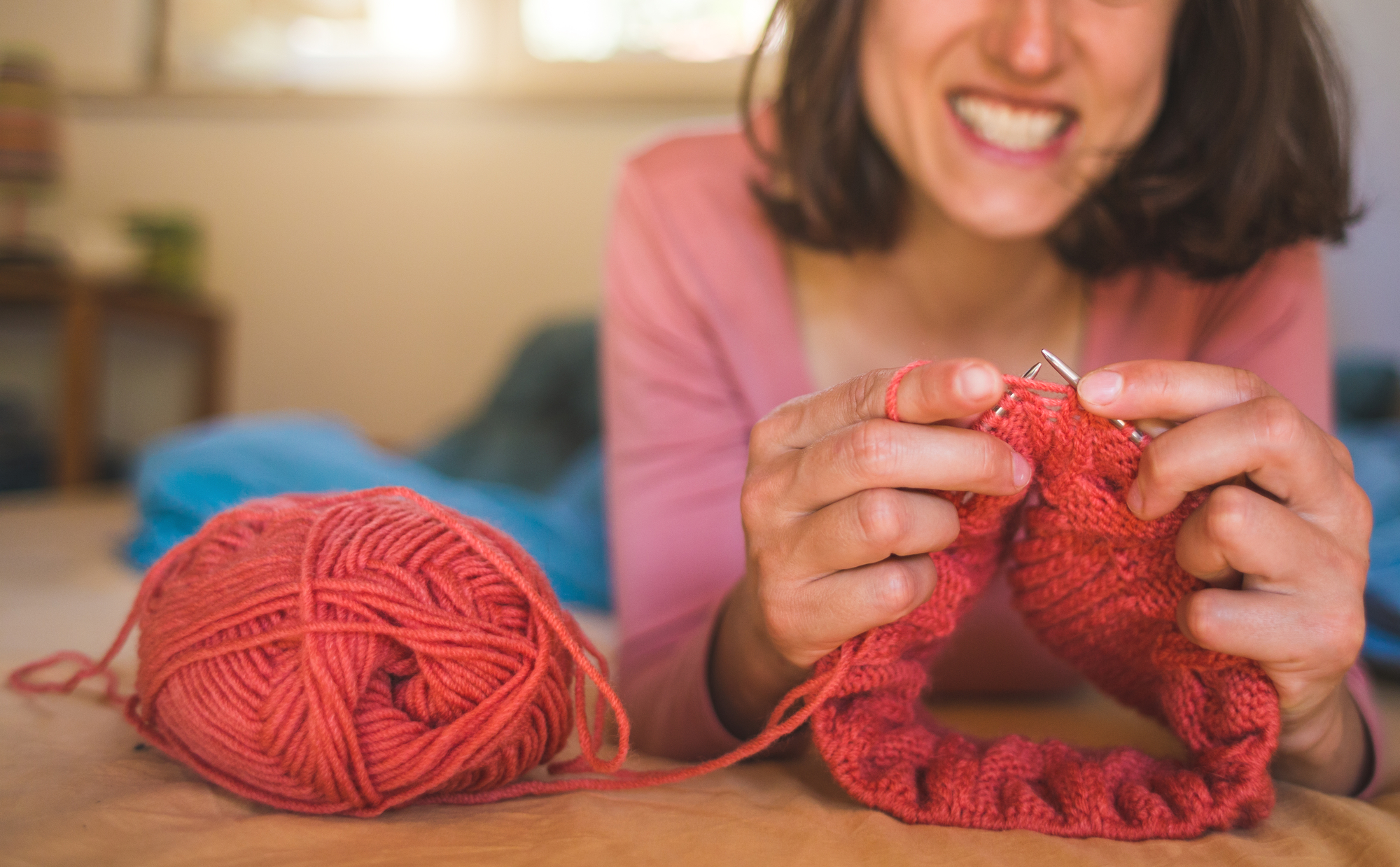 A person is smiling broadly while knitting with red yarn. They hold knitting needles and a partially finished piece, with the yarn ball placed on a flat surface. They are wearing a pink top, and the background appears to be a cozy indoor space.