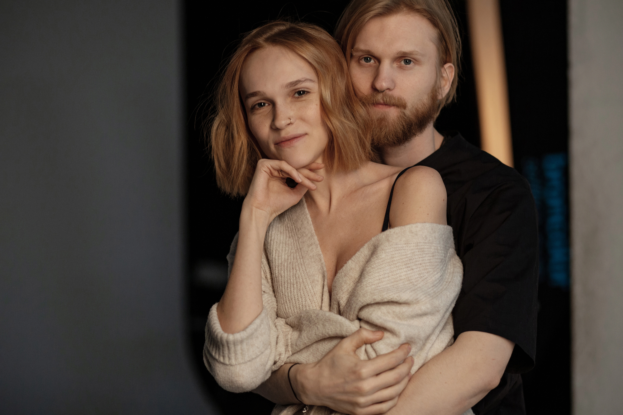 A woman with short blonde hair wrapped in a beige sweater is being embraced from behind by a man with medium-length blond hair and a beard. They both appear content and relaxed, standing against a dark background.