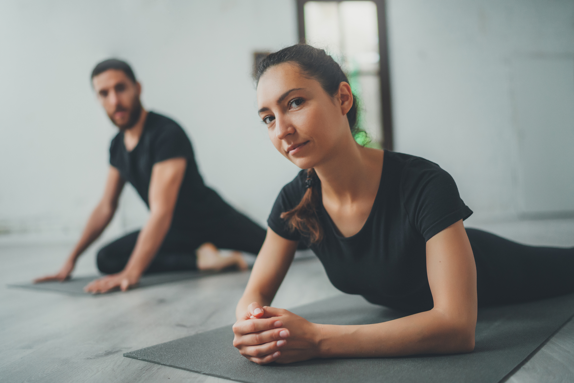 Two people wearing black workout clothing are doing yoga on mats indoors. The woman in the foreground is lying on her stomach with her hands clasped, while the man in the background is seated and stretching. The space appears calm and minimalistic.