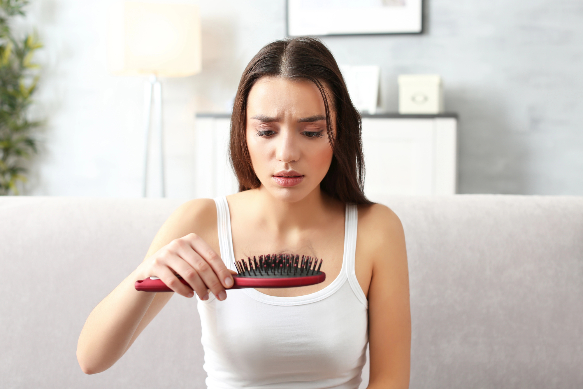 A young woman with long brown hair looks down at a hairbrush she's holding, visibly concerned. She is sitting on a light grey couch in a living room setting with a lamp and a shelf in the background.