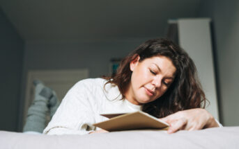 A person with long hair lies on a bed, propped up on their elbows, and reads a book or journal. They are wearing a cozy, white sweater and grey socks, with the background showing a modest, softly lit bedroom.