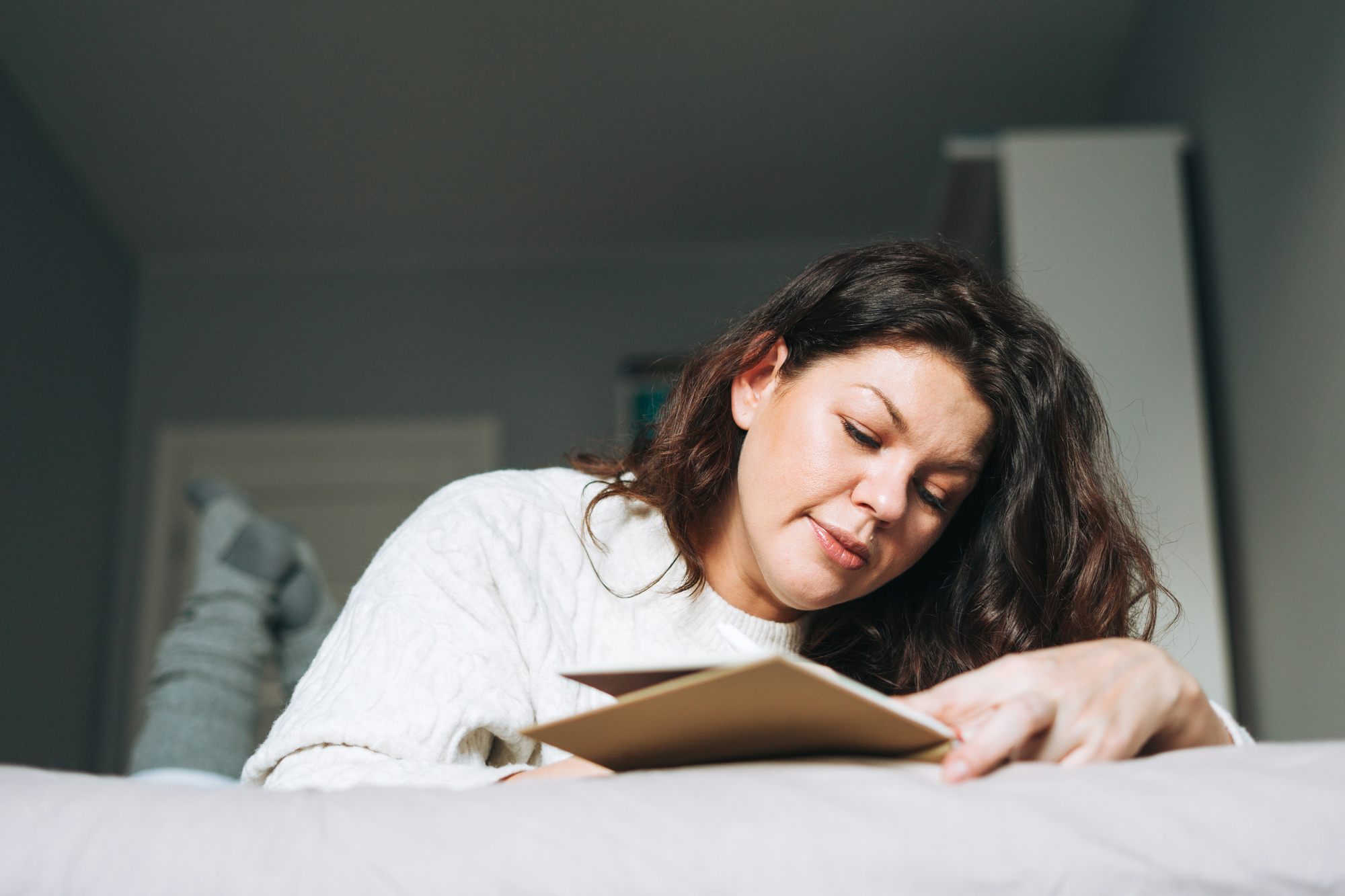 A person with long hair lies on a bed, propped up on their elbows, and reads a book or journal. They are wearing a cozy, white sweater and grey socks, with the background showing a modest, softly lit bedroom.