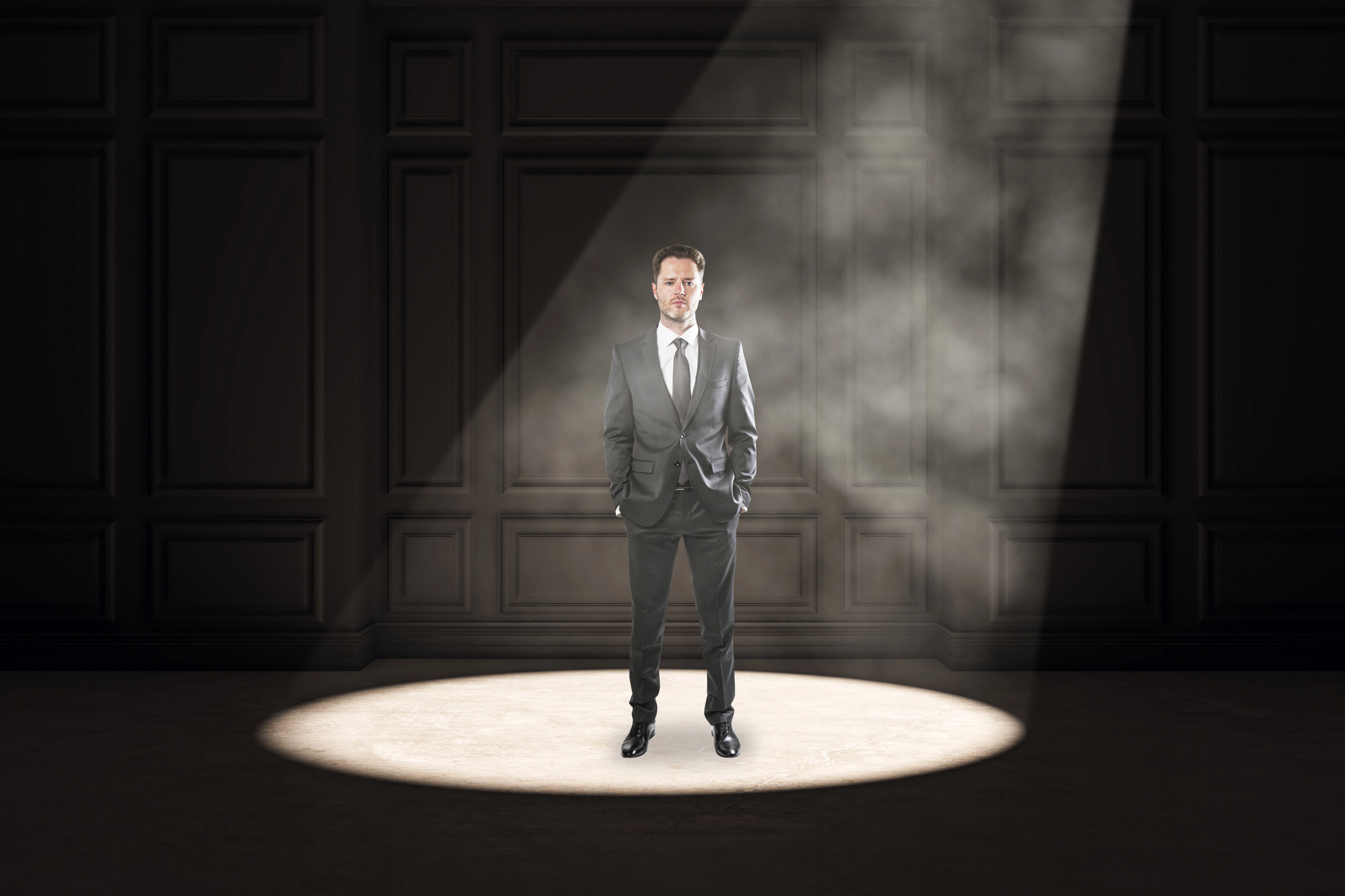 A man in a suit stands confidently under a spotlight in a dimly lit room with dark wood-paneled walls. His hands are in his pockets, and he is facing forward, casting a shadow behind him on the floor. A smoky haze is visible in the beam of light.