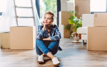 A woman in a plaid shirt and jeans sits on a wooden floor, surrounded by cardboard moving boxes. She rests her chin on her hand, gazing thoughtfully into the distance. The room has large windows, some sunlight, and a potted plant in the background.