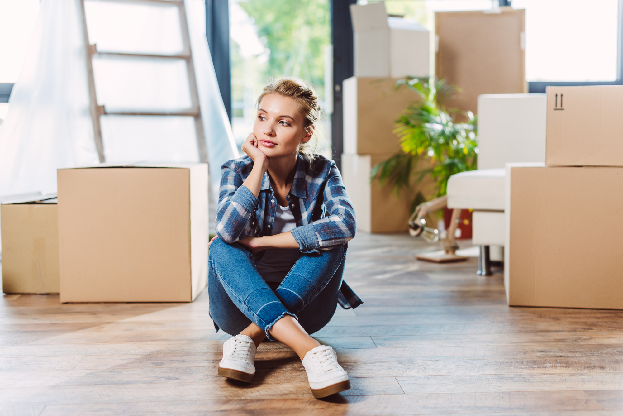 A woman in a plaid shirt and jeans sits on a wooden floor, surrounded by cardboard moving boxes. She rests her chin on her hand, gazing thoughtfully into the distance. The room has large windows, some sunlight, and a potted plant in the background.