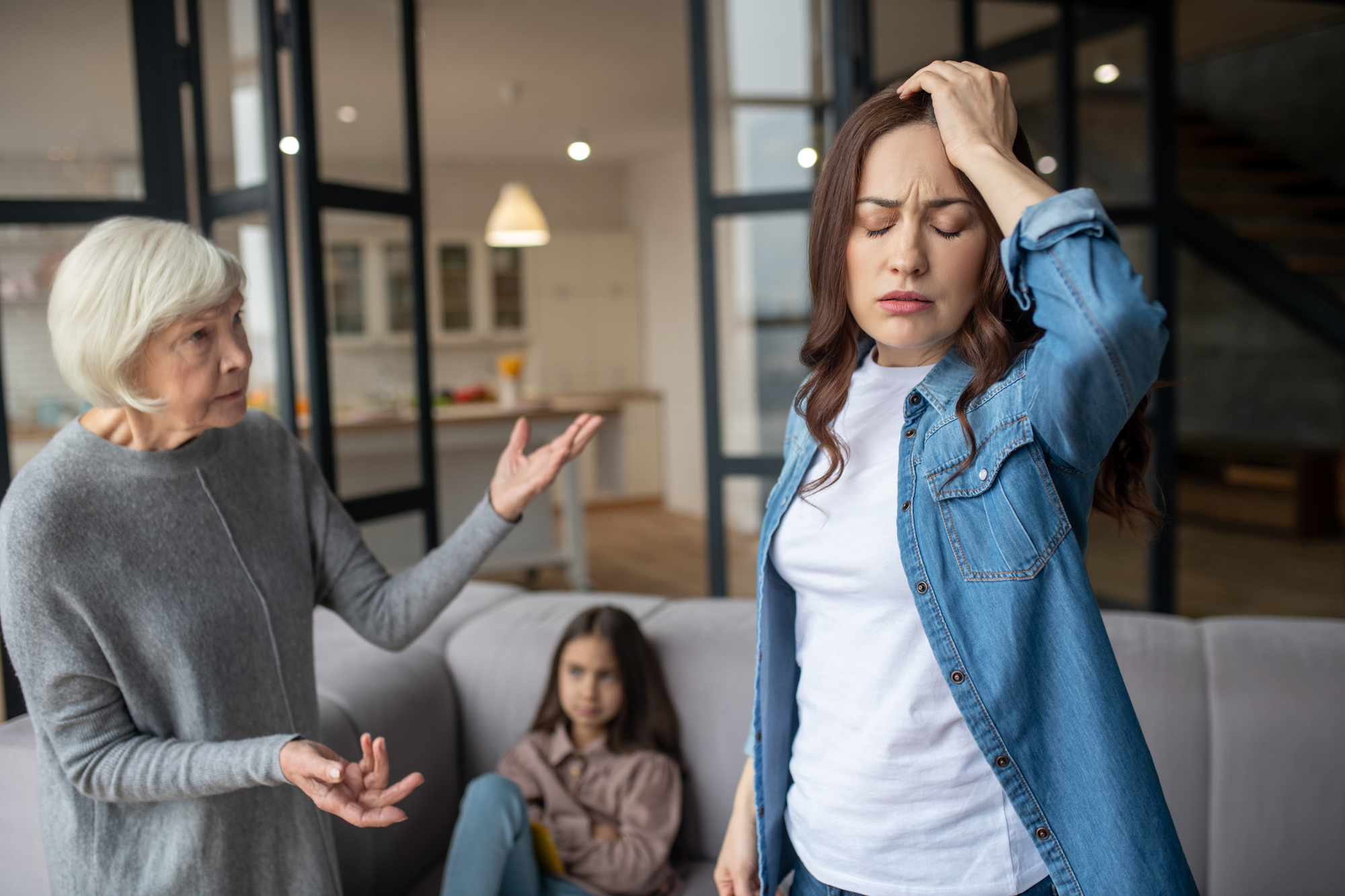 An elderly woman and a younger woman are having a heated discussion in a living room. The younger woman touches her head in frustration, while a young girl sits on a couch in the background, observing the scene with a concerned expression.