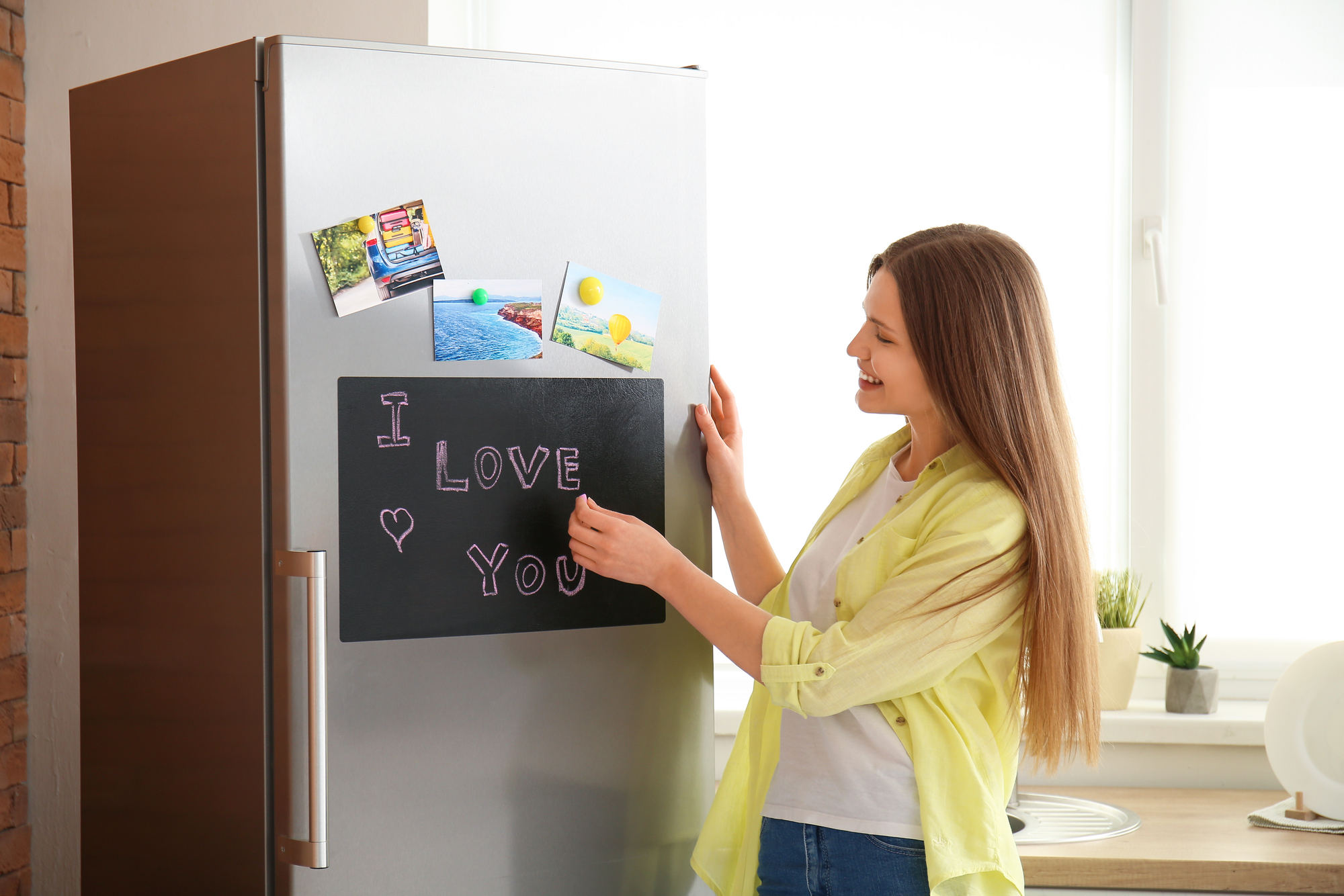 A woman with long brown hair, wearing a yellow shirt over a white top, smiles while arranging magnets and photos on a silver refrigerator. The fridge has a blackboard with "I love you" written on it in pink chalk. The background shows a bright kitchen.