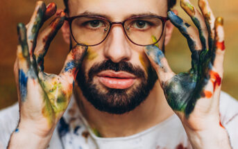 A bearded man with glasses and a serious expression holds his hands up to his face. His hands are covered in various colored paint smudges, and he wears a white shirt that is also stained with paint. The background is a neutral, blurred tone.