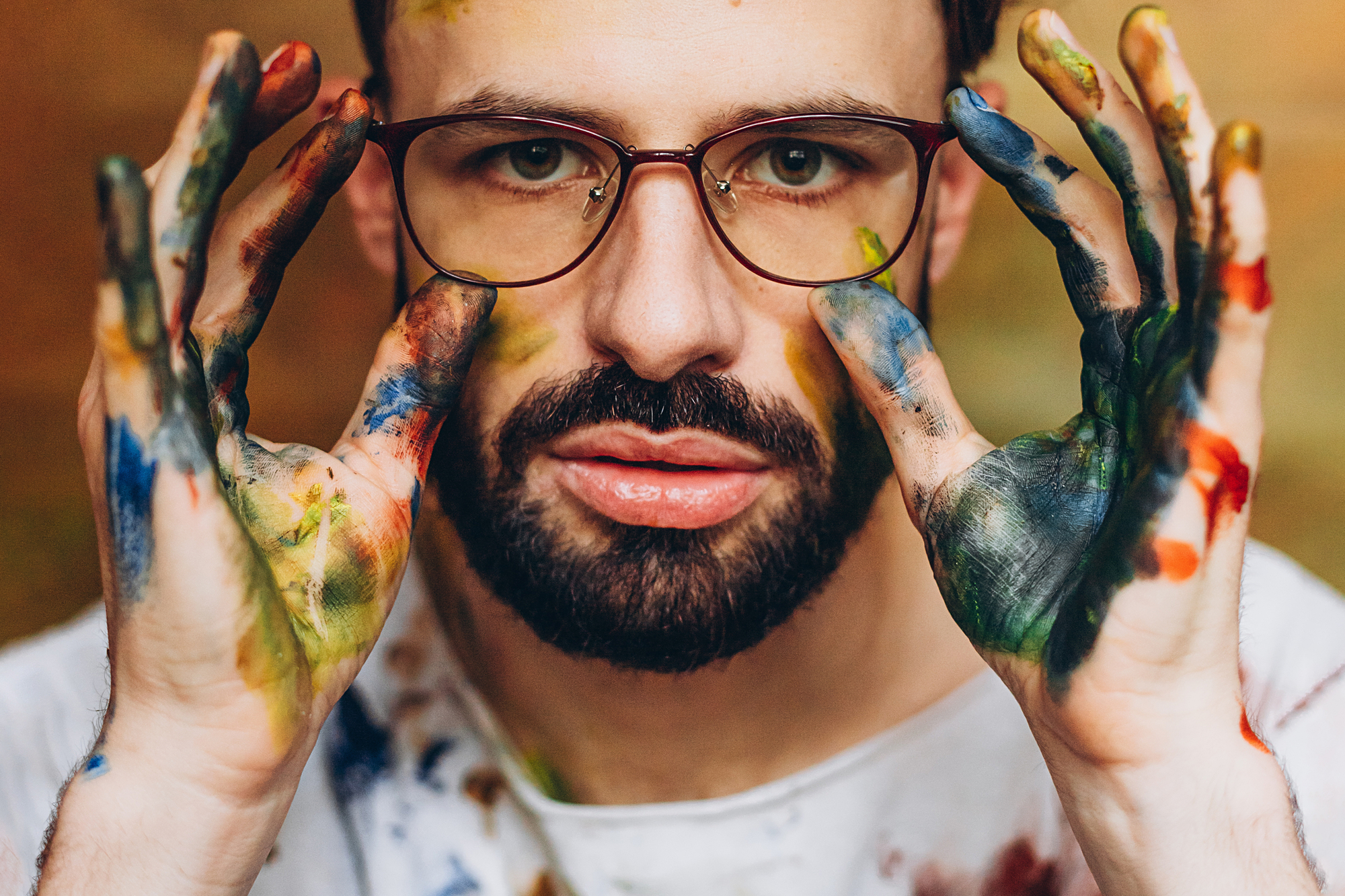 A bearded man with glasses and a serious expression holds his hands up to his face. His hands are covered in various colored paint smudges, and he wears a white shirt that is also stained with paint. The background is a neutral, blurred tone.