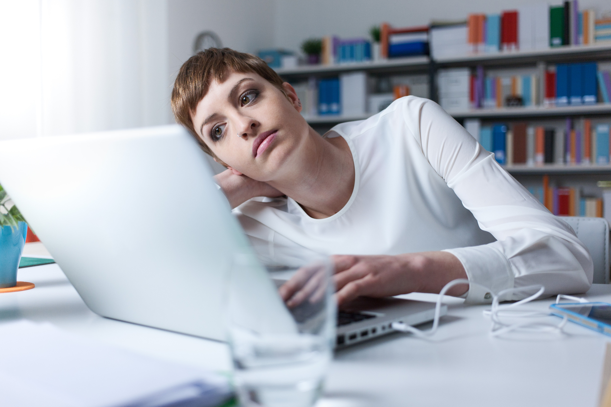 A person with short hair sits at a desk, resting their head on their hand while looking at a laptop screen. They appear tired or thoughtful. The background includes shelves filled with colorful binders and books. A glass and several documents are on the desk.