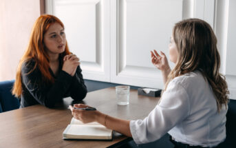 Two women are engaged in a serious conversation at a wooden table in an office setting. One woman with long red hair is listening carefully with hands clasped, while the other woman with light brown hair gestures while speaking. An open notebook and glass of water are on the table.