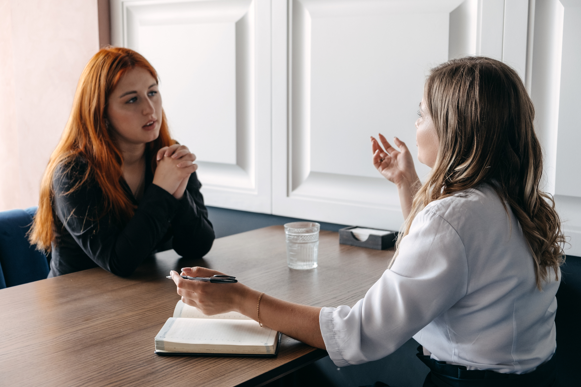 Two women are engaged in a serious conversation at a wooden table in an office setting. One woman with long red hair is listening carefully with hands clasped, while the other woman with light brown hair gestures while speaking. An open notebook and glass of water are on the table.