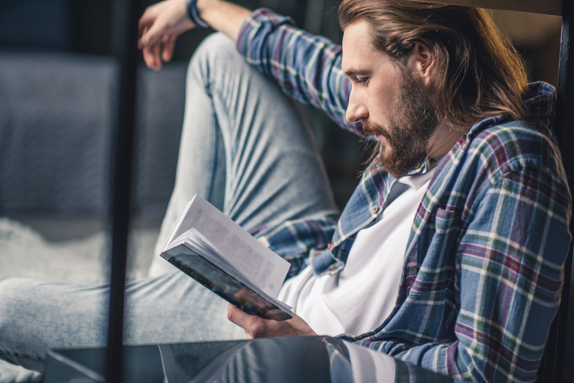 A man with long hair and a beard is sitting on the floor, leaning against a piece of furniture. He is wearing a casual plaid shirt over a white T-shirt and light blue jeans, and he’s absorbed in reading a book. The setting appears to be a cozy indoor space.