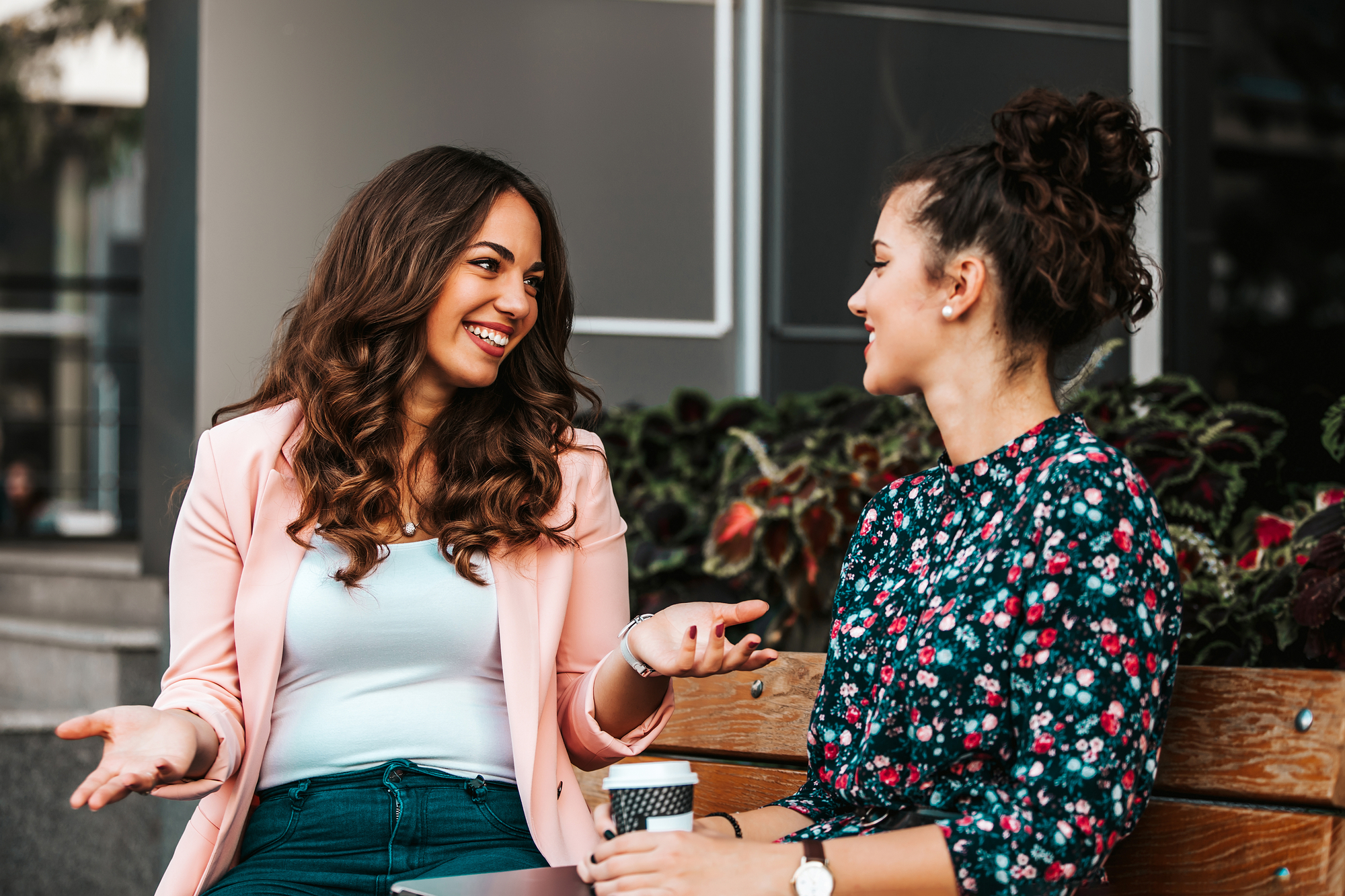 Two women sitting on a bench outdoors, engaged in conversation. One woman, with wavy hair, wears a light pink blazer and white top, gesturing with her hands. The other woman, with a bun, wears a floral blouse and holds a coffee cup. Green plants are in the background.